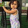 Catalina is registered to the contest to win money with this photo: toddler, child, curly_hair, romper, pastel_colors, wicker_chair, hand, outdoor, green_carpet, bracelet, expression, portrait, standing, furniture, cute, person, young_child, summer_clothing, natural_light, curiosity