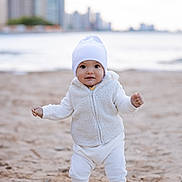 Ostap is registered to the contest to win money with this photo: baby, beach, beanie, casual, child, cityscape, cute, daytime, footwear, infant, outdoor, portrait, sand, smiling, standing, urban_background, water, white_clothing, winter_clothes, young_child