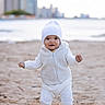 baby, beach, beanie, casual, child, cityscape, cute, daytime, footwear, infant, outdoor, portrait, sand, smiling, standing, urban_background, water, white_clothing, winter_clothes, young_child