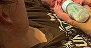 Victoria is registered to the contest to win money with this photo: baby, bottle, feeding, infant, person, adult, brown_shirt, text, hand, face, sleepy, indoor, closeup, holding, cuddle, skin, hair, relaxation, comfort, blanket
