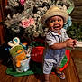 toddler, child, smiling, hat, romper, flip_flops, wooden_floor, christmas_tree, holiday, decorations, toys, stuffed_animal, bucket, ribbon, flower, indoor, happy, person, clapping, festive