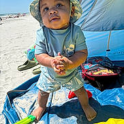 Omari is registered to the contest to win money with this photo: toddler, child, beach, sand, sun_hat, swimwear, blue_shirt, shorts, sunny, outdoor, barefoot, playing, sand_toys, tent, backpack, water_toys, happy, summer, vacation, portrait