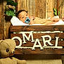 Omari is registered to the contest to win money with this photo: baby, pacifier, wooden_crate, name_letters, teddy_bear, greenery, wooden_floor, wooden_wall, blanket, infant, indoor, soft_toy, cute, child, relaxing, decor, portrait, nursery, cozy, resting