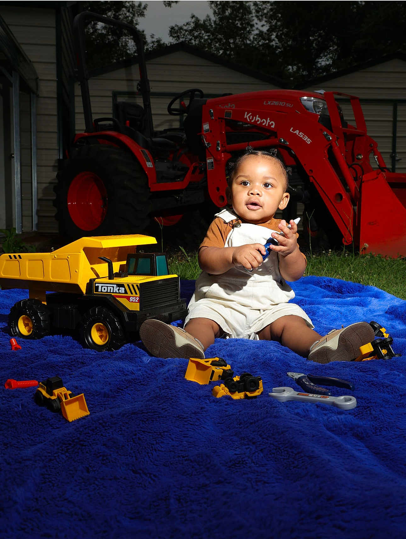 Omari is registered to the contest to win money with this photo: toddler, child, toy, toy_truck, construction_toy, blue_blanket, outdoor, grass, tractor, red_tractor, shed, play, smiling, young_child, sitting, sunlight, tools, yellow_toy, person, cute