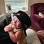 baby, child, cap, hat, smile, indoors, person, face, hand, arm, window, blind, furniture, couch, pillow, blanket, skin, clothing, holding, infant