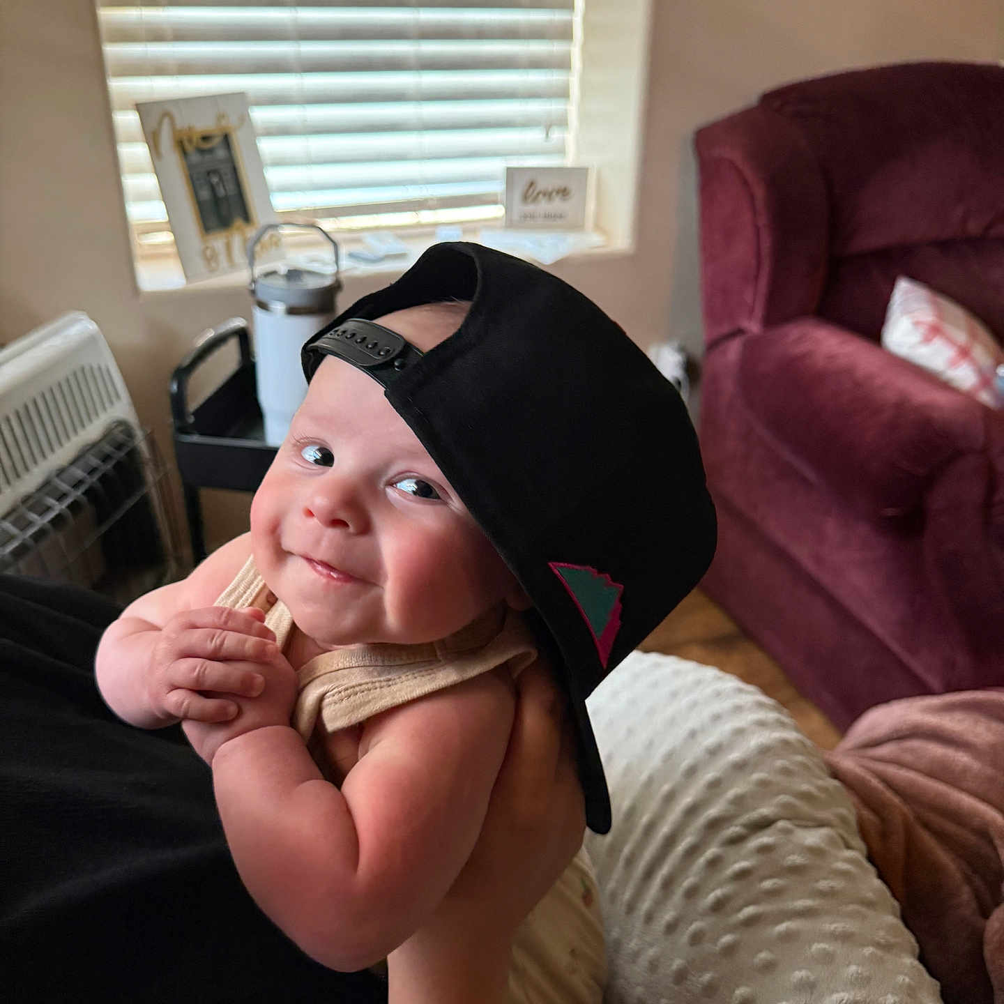 Asher is registered to the contest to win money with this photo: arm, baby, blanket, blind, cap, child, clothing, couch, face, furniture, hand, hat, holding, indoors, infant, person, pillow, skin, smile, window