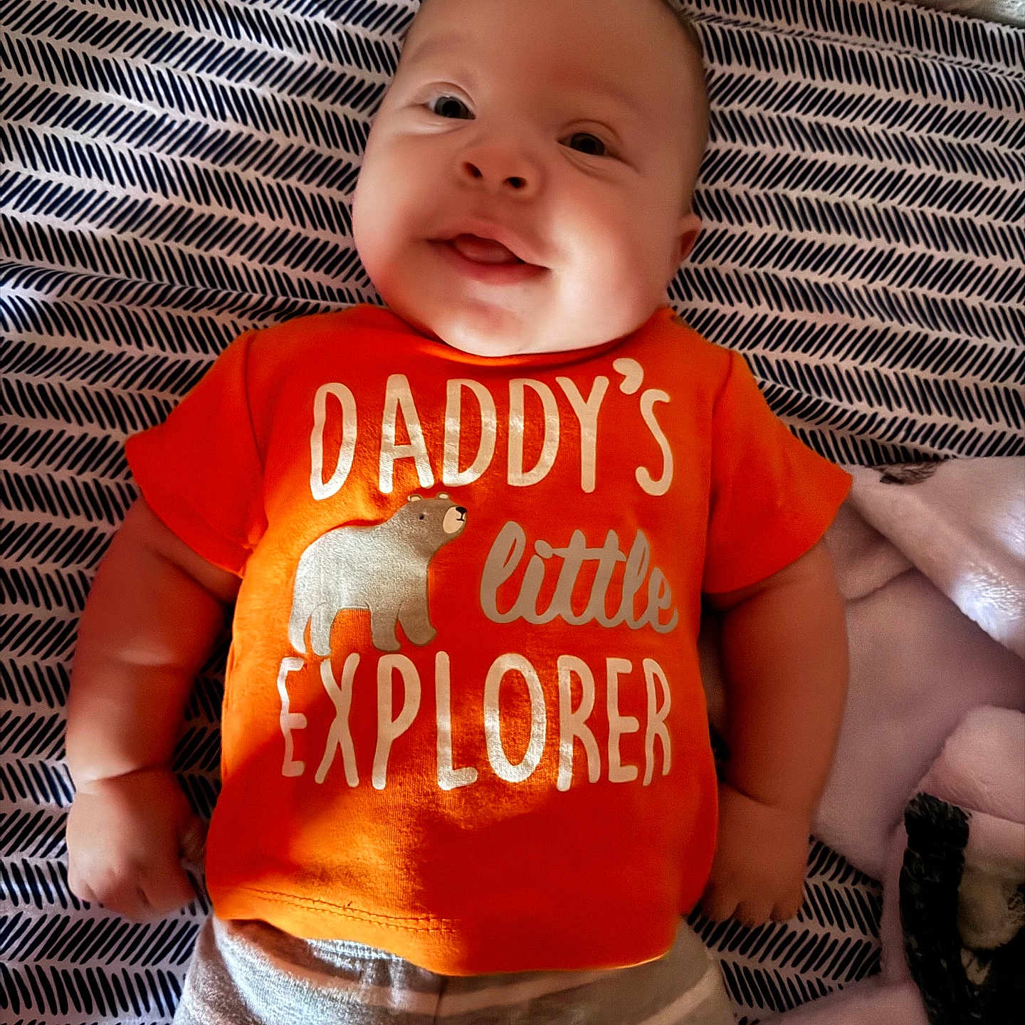 Liam joined the competition — help win amazing prizes! baby, blanket, child, clothing, cute, explorer, face, happy, indoors, infant, lying_down, orange_shirt, pattern, portrait, smile, soft_lighting, striped_pants, text_on_clothing, toddler, young_child