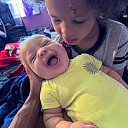 Liam is registered to the contest to win money with this photo: baby, child, smiling, laughing, curly_hair, yellow_onesie, indoor, bedroom, blanket, hand, tender, holding, person, candid, family, happy, infant, sibling, portrait, cozy