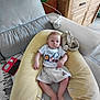 baby, child, couch, pillow, cushion, furniture, indoor, person, toddler, clothing, shorts, tshirt, wooden_cabinet, floor_tiles, electronic_device, power_strip, blanket, rug, relaxing, home