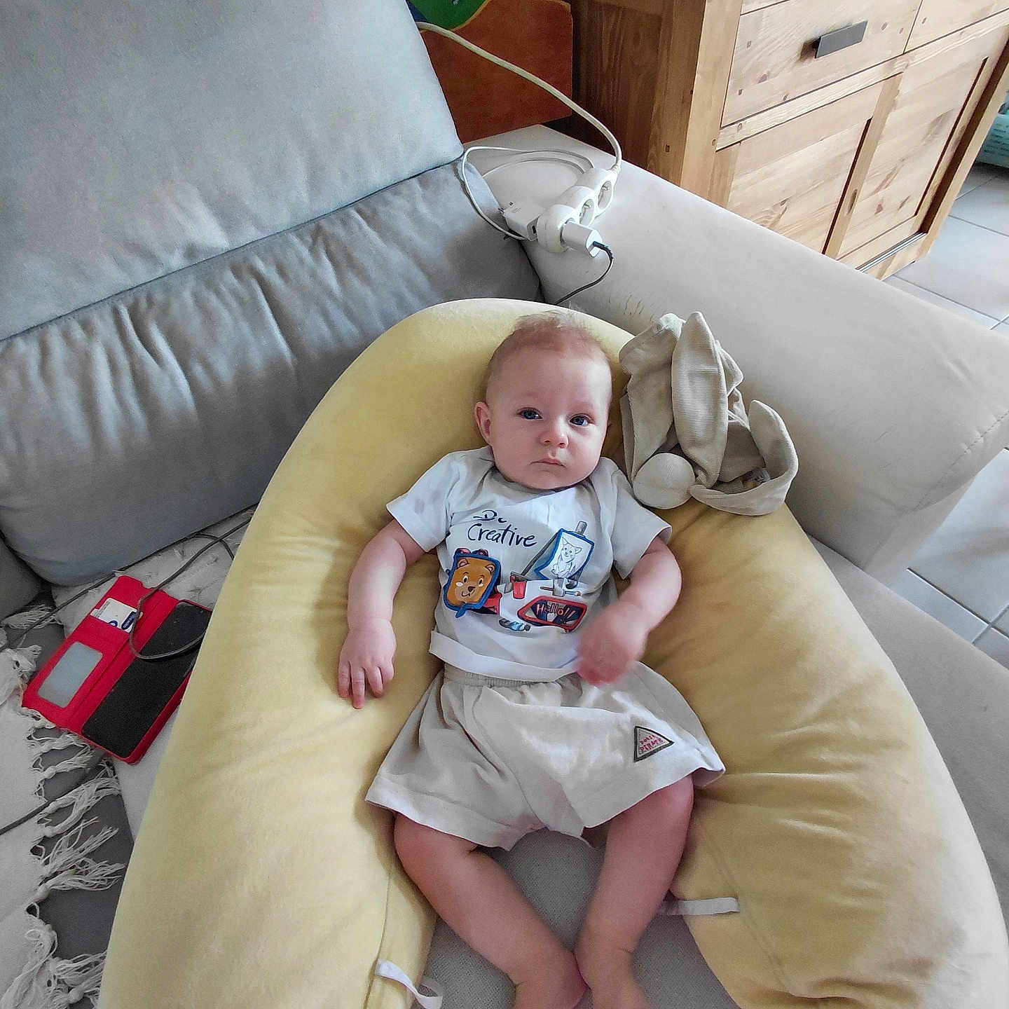 Tiago participe au concours pour gagner de l'argent avec cette photo : baby, blanket, child, clothing, couch, cushion, electronic_device, floor_tiles, furniture, home, indoor, person, pillow, power_strip, relaxing, rug, shorts, toddler, tshirt, wooden_cabinet