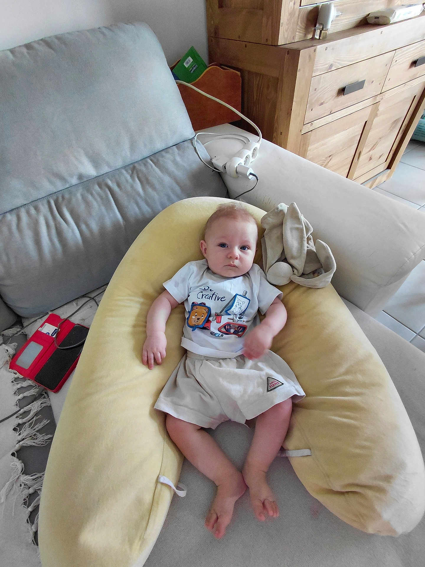 Tiago participe au concours pour gagner de l'argent avec cette photo : baby, child, couch, pillow, cushion, furniture, indoor, person, toddler, clothing, shorts, tshirt, wooden_cabinet, floor_tiles, electronic_device, power_strip, blanket, rug, relaxing, home