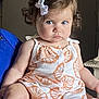 Kinsley joined the competition — help win amazing prizes! baby, book, child, cross, curly_hair, decor, footwear, furniture, hand, human, indoor, orange_pattern, person, romper, serious_expression, sitting, skin, table, wall, white_bow
