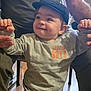 Francescu-Maria participe au concours pour gagner de l'argent avec cette photo : baby, cap, casual_clothing, child, face, family, floor, grey_shirt, hands, happy, headwear, human, indoor, orange_text, pants, person, portrait, smiling, support, toddler
