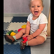 Camil participe au concours pour gagner de l'argent avec cette photo : toddler, child, laughing, indoor, toy, foam_mats, white_shirt, beige_shorts, brown_shoes, curly_hair, floor, happy, playtime, person, smile, furniture, wooden, purple_wall, sitting, young_child