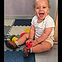 toddler, child, laughing, indoor, toy, foam_mats, white_shirt, beige_shorts, brown_shoes, curly_hair, floor, happy, playtime, person, smile, furniture, wooden, purple_wall, sitting, young_child