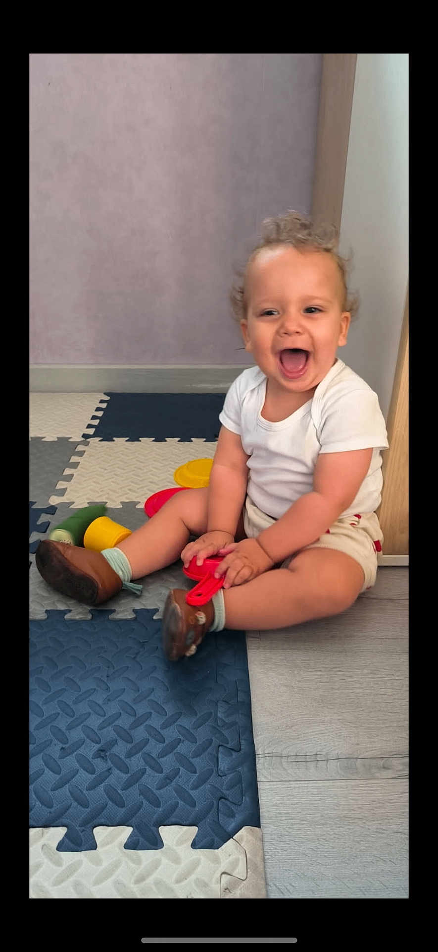 Camil participe au concours pour gagner de l'argent avec cette photo : toddler, child, laughing, indoor, toy, foam_mats, white_shirt, beige_shorts, brown_shoes, curly_hair, floor, happy, playtime, person, smile, furniture, wooden, purple_wall, sitting, young_child