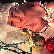 Odessa is registered to the contest to win money with this photo: baby, infant, santa_hat, christmas_lights, ornaments, holiday, festive, blanket, soft_texture, cute, surprised_expression, indoors, decorations, holiday_season, red, gold, lighting, celebration, child, festivity