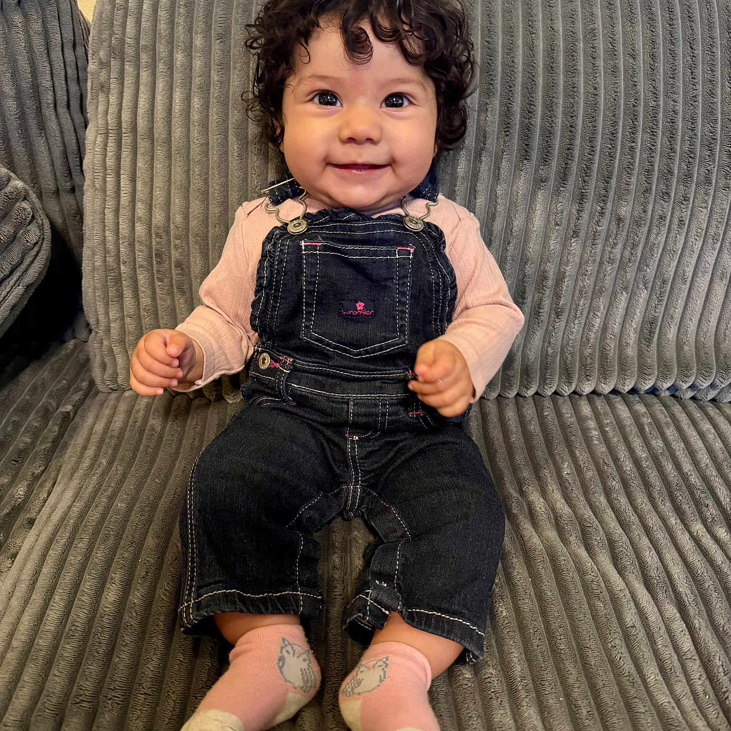 Demri is registered to the contest to win money with this photo: animal_socks, baby, child, couch, cozy, curly_hair, cute, denim_overalls, gray_couch, happy, indoors, person, pink_bow, pink_shirt, pink_socks, portrait, sitting, smiling, textured_couch, young_child