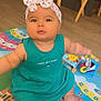 baby, child, headband, bow, dress, toy, play_mat, floor, wooden_chair, indoor, colorful, alphabet, infant, sitting, cute, person, foot, hand, face, curious