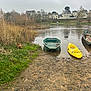 dog, boat, river, water, grass, mud, trees, houses, cloudy_sky, nature, outdoor, village, dock, paddle, yellow_kayak, green_boat, brown_dog, reeds, calm, overcast