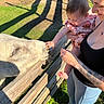adult, animal, baby, bridge, curious, dress, family, farm, goat, grass, held, infant, outdoor, parent, portrait, reaching, sunlight, tattoo, wooden_fence, zoo