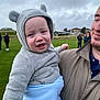 baby, child, man, outdoor, grass, cloudy_sky, hoodie, blanket, smile, people, casual_clothing, portrait, holding, cute, warm_clothing, park, family, happy, infant, headwear