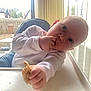 Nate is registered to the contest to win money with this photo: baby, high_chair, food, snack, hand, face, window, curtain, indoor, child, messy, white_clothing, table, fence, outdoor, daylight, skin, head, eating, person