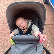 Nate is registered to the contest to win money with this photo: baby, stroller, smiling, child, toy, outdoor, pavement, car, blue, gray, clothing, happy, infant, cute, seatbelt, person, head, hand, playful, transport