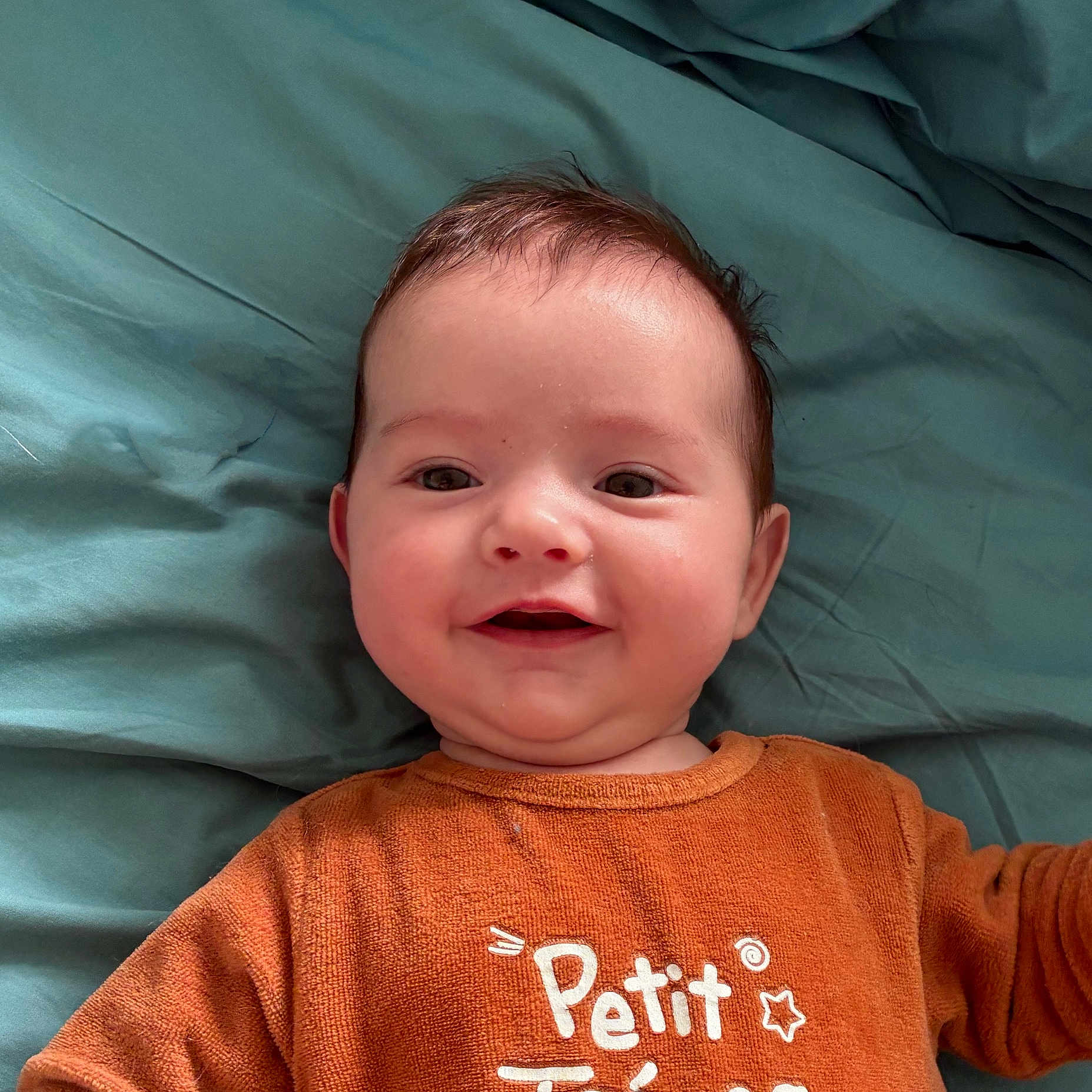 Mahé a rejoint le concours — aidez-le/la à gagner de superbes lots ! baby, smiling, infant, orange_clothing, lying_down, blanket, face, child, cute, happy, portrait, head, skin, young_child, eyes, mouth, cheeks, hair, text_on_clothing, indoors