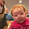 toddler, child, glasses, pink_shirt, smiling_face, curious_expression, woman, mask, man, blue_shirt, wooden_chair, indoor, ceiling_lights, people, portrait, close_up, seated, casual_clothing, hair, face