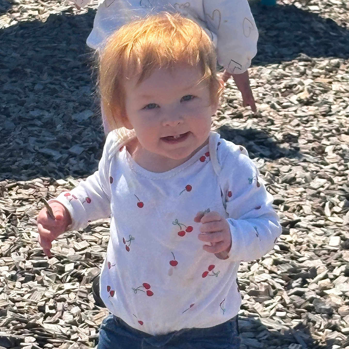 Nora is registered to the contest to win money with this photo: toddler, child, playground, woodchips, stick, white_shirt, cherry_pattern, red_hair, smiling, outdoor, sunlight, casual_clothing, hoodie, heart_pattern, person, two_children, nature, daylight, jeans, happy