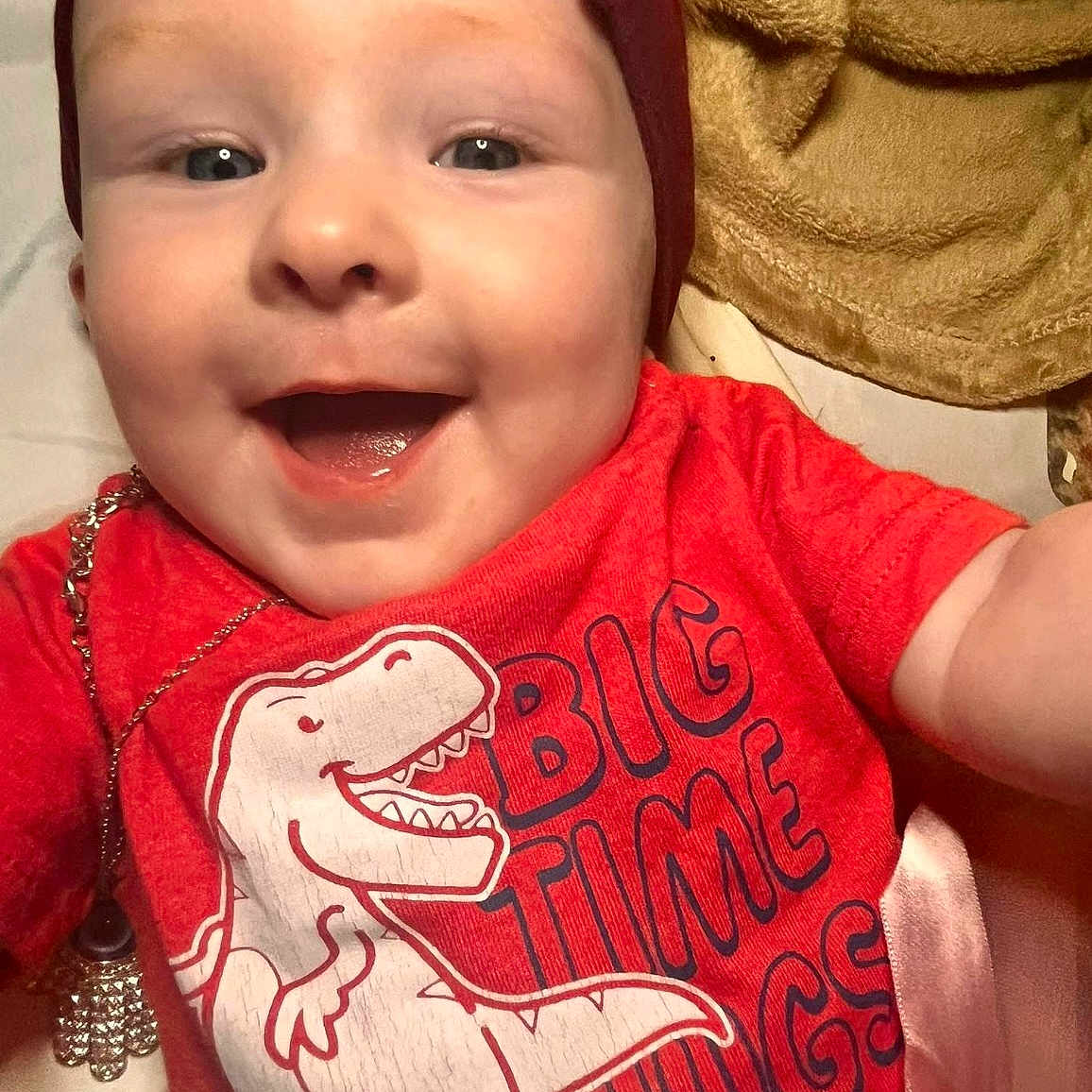Nora joined the competition — help win amazing prizes! baby, blanket, cheerful, child, closeup, clothing, cozy, cute, dinosaur_graphic, face, happy, headwear, indoor, infant, person, portrait, red_shirt, smiling, text_on_clothing, toddler