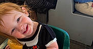 Nora is registered to the contest to win money with this photo: toddler, child, smile, bread, food, high_chair, restaurant, table, people, indoor, red_hair, clothing, black_top, red_skirt, cute, happy, dining, family, person, seat