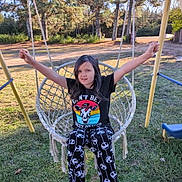 Alana is registered to the contest to win money with this photo: arms_raised, black_tshirt, casual_clothing, child, daylight, footwear, girl, graphic_tshirt, grass, happy, nature, outdoor, pants, play, playground, recreation, skull_pattern, smiling, swing, trees