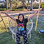 arms_raised, black_tshirt, casual_clothing, child, daylight, footwear, girl, graphic_tshirt, grass, happy, nature, outdoor, pants, play, playground, recreation, skull_pattern, smiling, swing, trees