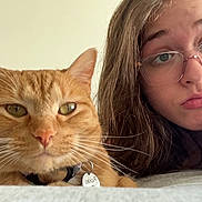 Pickles joined the competition — help win amazing prizes! cat, orange_tabby, person, glasses, bed, whiskers, face, pout, fur, collar, indoor, closeup, pet, young_adult, hair, expression, home, relaxing, animal, portrait