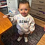 Mckenzie is registered to the contest to win money with this photo: toddler, child, smiling, standing, curly_hair, indoor, sweatshirt, shorts, shoes, table, rug, furniture, drawer, cabinet, earrings, wooden_floor, patterned_rug, holding, bright_eyes, happy