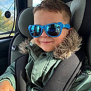 Arlo is registered to the contest to win money with this photo: child, car_seat, sunglasses, blue_sunglasses, jacket, fur_hood, smile, seat_belt, car_interior, reflection, hand, window, sunlight, casual_clothing, young_child, portrait, happy, kid, person, indoors