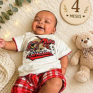Miguel is registered to the contest to win money with this photo: baby, infant, smiling, dinosaur_shirt, red_plaid_shorts, teddy_bear, wooden_milestone, 4_months, fairy_lights, cream_blanket, cozy, cute, child, portrait, happy, lying_down, soft_texture, plush_toy, milestone_sign, home