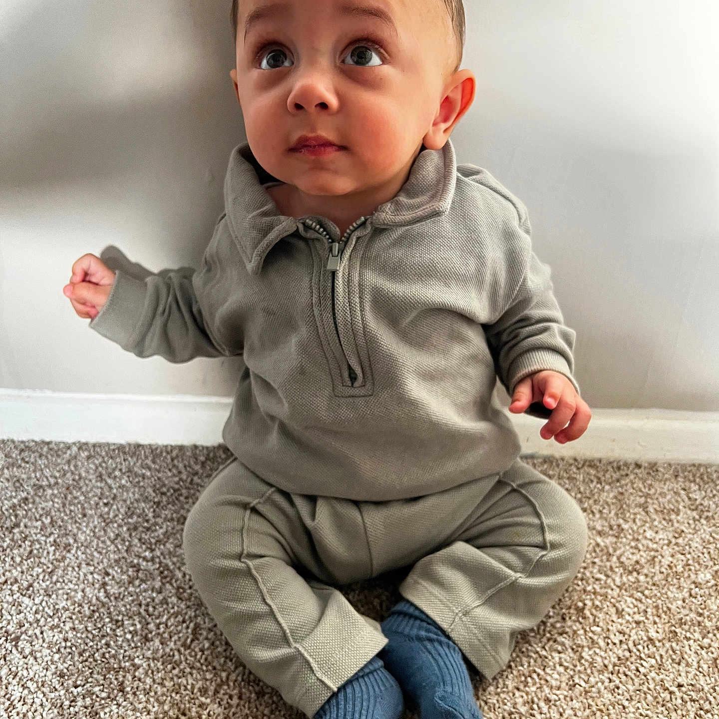 Amaari is registered to the contest to win money with this photo: baby, blue_socks, carpet, casual, child, curious, cute, expression, face, floor, gray_clothing, head, indoor, person, portrait, sitting, small_hands, toddler, wall, young_child