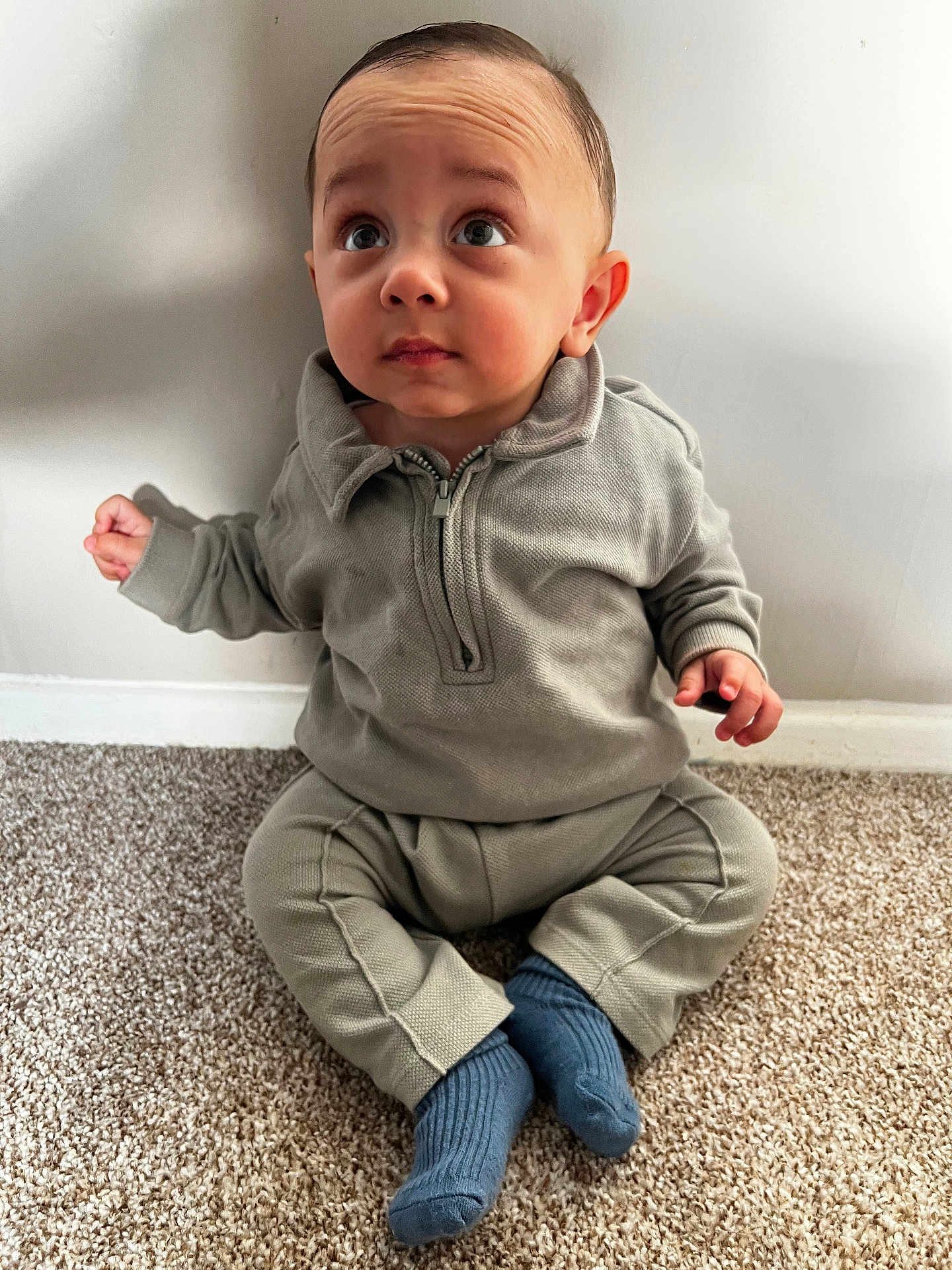 Amaari is registered to the contest to win money with this photo: toddler, child, baby, sitting, gray_clothing, blue_socks, carpet, indoor, curious, expression, face, head, person, wall, floor, casual, young_child, small_hands, cute, portrait