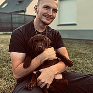 Arés a rejoint le concours — aidez-le/la à gagner de superbes lots ! young_man, puppy, dog, chocolate_labrador, grass, outdoor, house, smiling, sitting, bracelet, chain_necklace, black_tshirt, casual_wear, happy, pet, portrait, daylight, backyard, fence, relaxed