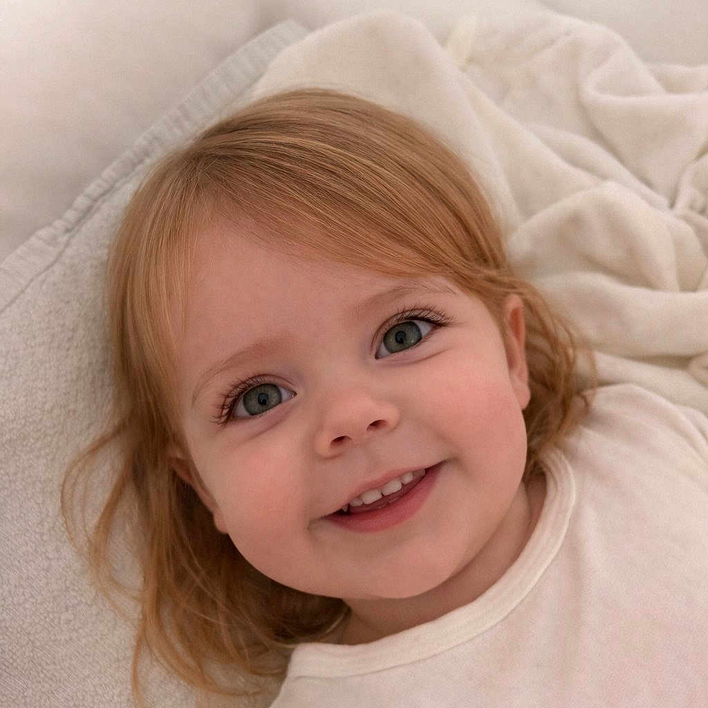 Naya a rejoint le concours — aidez-le/la à gagner de superbes lots ! toddler, child, smile, face, blond_hair, blue_eyes, white_clothing, blanket, soft_texture, indoors, cozy, happy, portrait, cute, young_child, laying_down, close_up, innocence, comfort, gentle_lighting