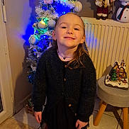 Mélyna participe au concours pour gagner de l'argent avec cette photo : child, girl, christmas_tree, holiday_decorations, boots, black_cardigan, smiling, indoor, floor_tiles, radiator, santa_figurine, snowman_figurine, ornaments, lights, festive, necklace, skirt, carpet, stool, home