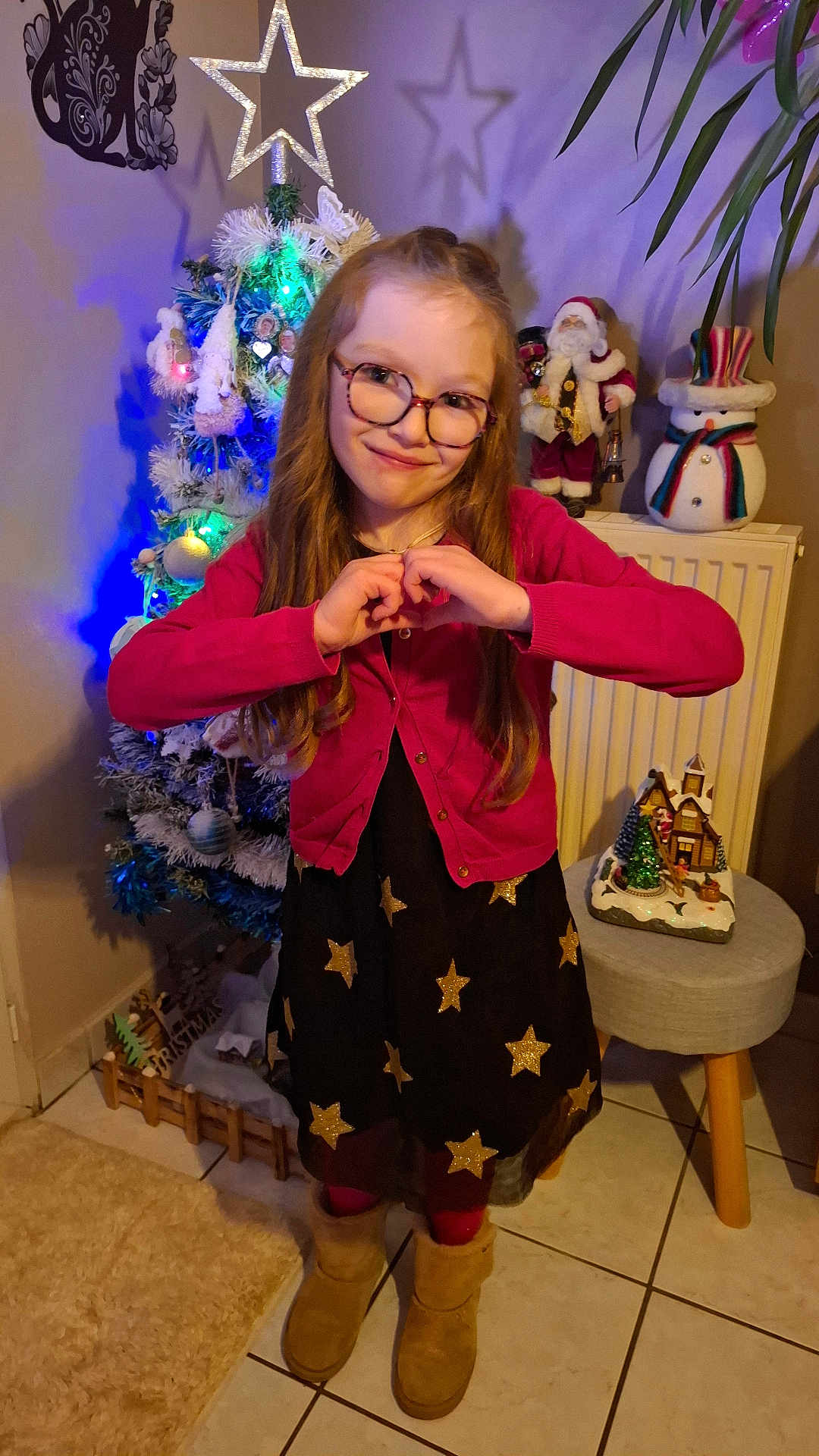 Maëlle participe au concours pour gagner de l'argent avec cette photo : child, girl, glasses, dress, stars, cardigan, boots, christmas_tree, holiday_decor, snowman, santa_figurine, indoor, smile, pose, floor_tiles, plant, furniture, festive, warm_lighting, home