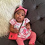 baby, child, infant, pink_clothing, headband, purse, couch, plush_pillow, floral_pattern, tufted_furniture, sitting, cute, accessory, indoors, portrait, smiling, happy, toddler, young_child, cozy