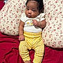 baby, infant, child, bow, cushion, floral_pattern, yellow_pants, white_top, cute, smiling, relaxed, indoors, portrait, clothing, footwear, skin, hair, bracelet, red_bedspread, comfort