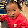 background, candid, casual_clothing, chairs, child, curly_hair, earrings, expression, face, indoor, people, person, pigtails, red_sweatshirt, seated, stroller, toddler, waiting, waiting_room, wood_floor