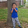 blue_shirt, brick_wall, casual_clothing, child, curly_hair, daytime, dinosaur_pants, grass, happy, house, leaves, nature, outdoor, playful, shoes, smiling, standing, tree, window, yard