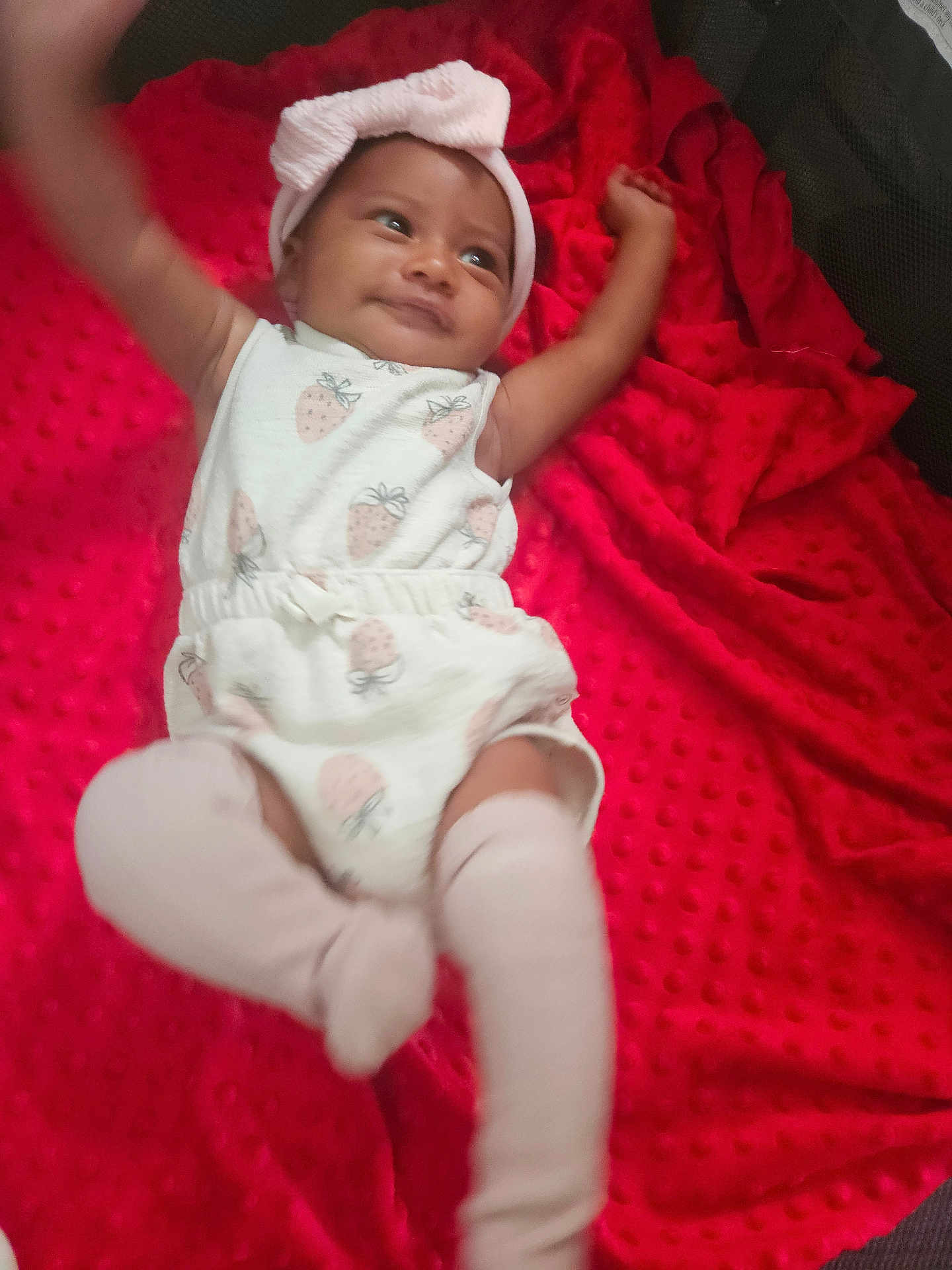 L'Eylah is registered to the contest to win money with this photo: baby, infant, child, smiling, headband, white_clothing, strawberry_pattern, red_blanket, blanket, soft_texture, lying_down, happy, cute, indoors, skin, arms_up, legs_up, sock, headwear, playful